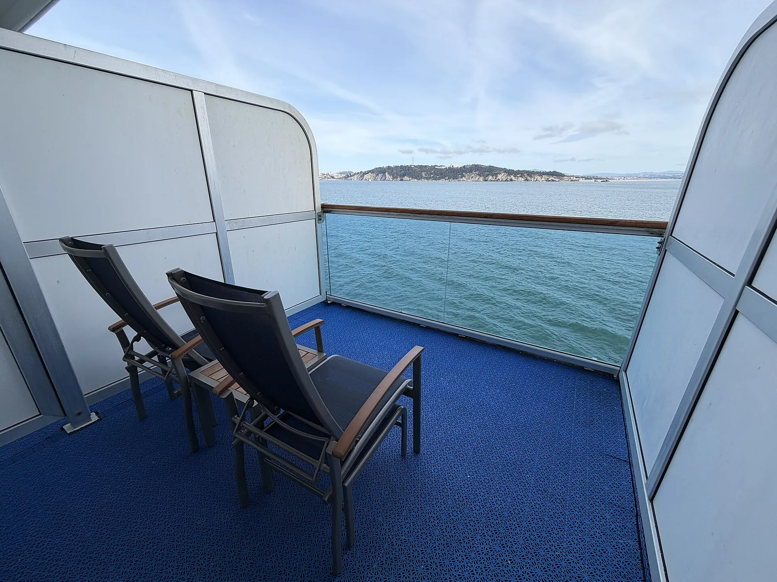 Two chairs on a private stateroom balcony with glass railings, looking out over blue water and a green headland
